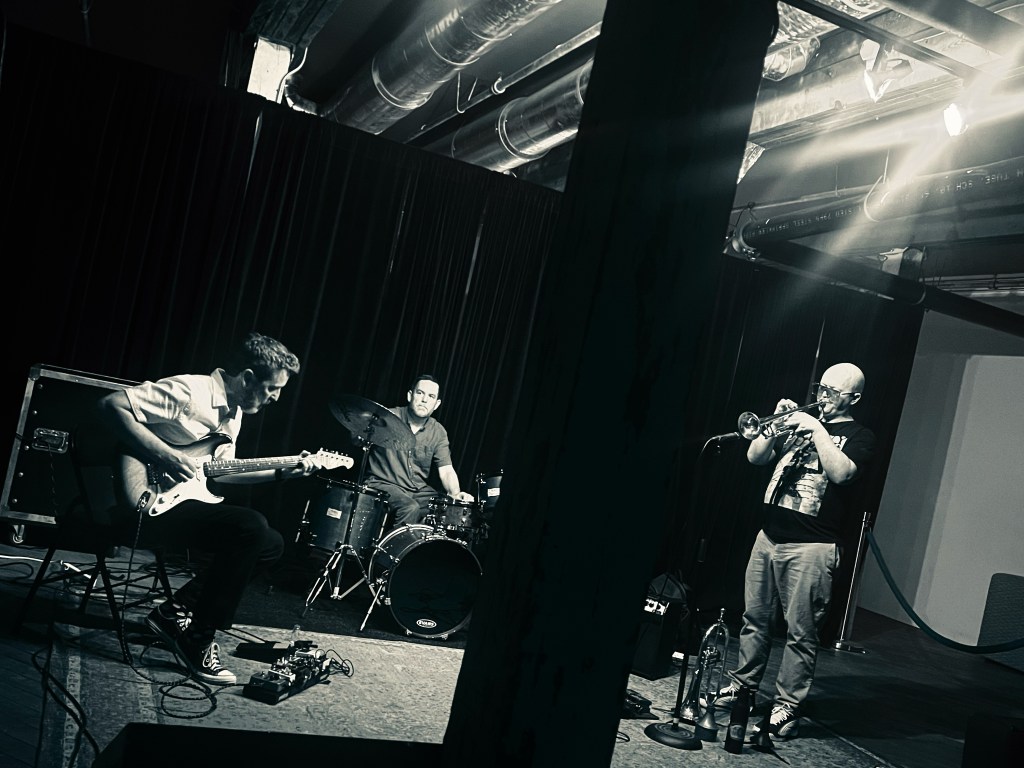 A guitarist, drummer, and trumpet playing in a dark club atmosphere